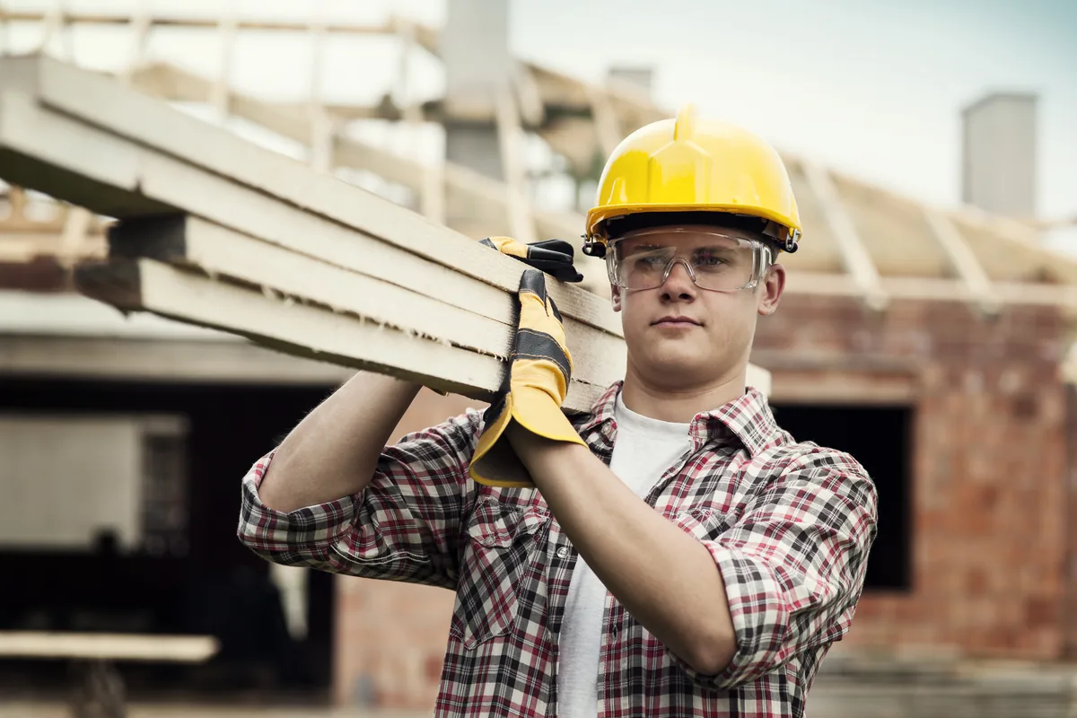 Bauarbeiter mit gelbem Schutzhelm und Schutzbrille trägt Holzbretter auf der Schulter, im Hintergrund ein Rohbau mit Dachstuhl